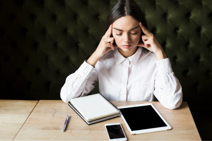 Person at a desk with notebook, tablet, and phone while pressing temples to show tracking of headache symptoms and daily habits