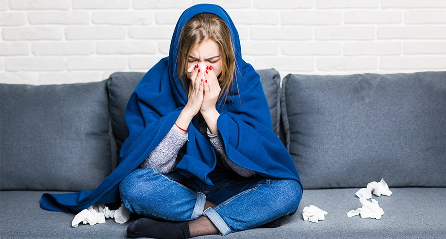 A person wrapped in a blanket, sneezing into a tissue while experiencing flu symptoms at home.